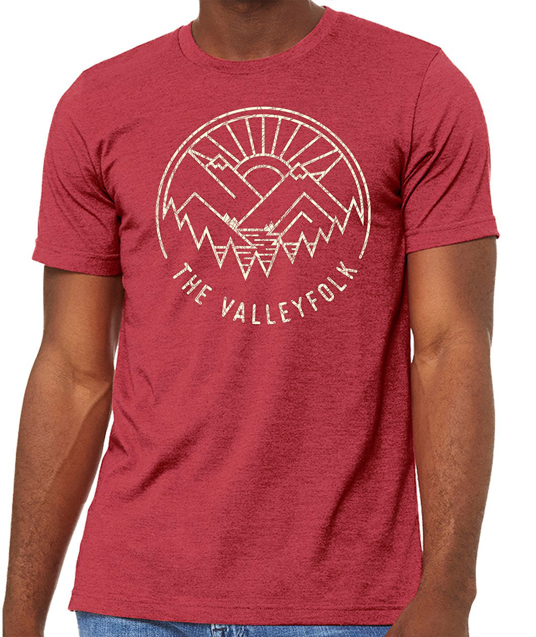 A red shirt with a distressed circular logo of a white outline of mountains, a lake, trees, and a sun rising. “The Valleyfolk” is arched at the bottom in white sans serif font - from the Valleyfolk