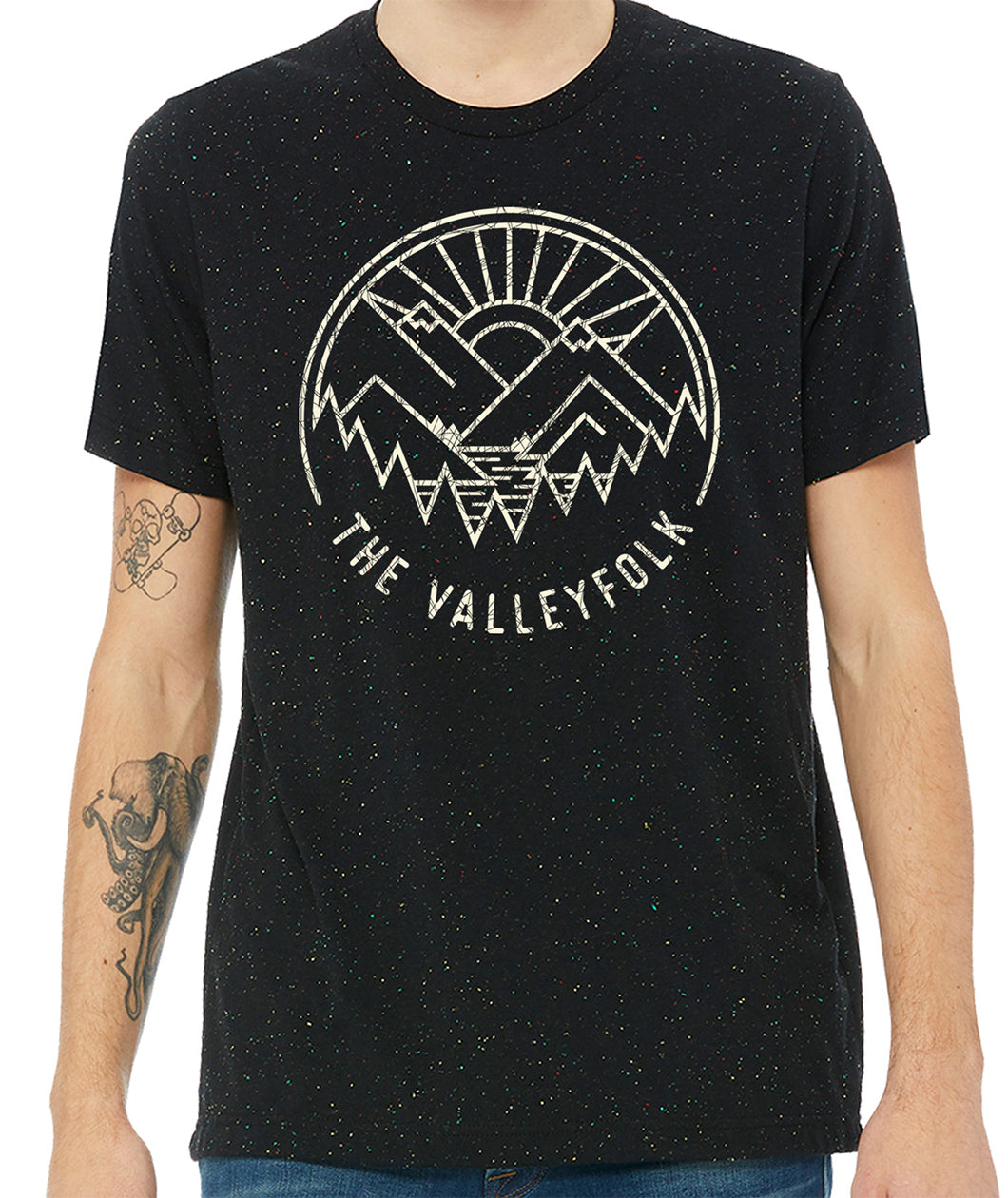 A black shirt with red yellow and blue speckles with a distressed circular logo of a white outline of mountains, a lake, trees, and a sun rising. “The Valleyfolk” is arched at the bottom in white sans serif font - from the Valleyfolk