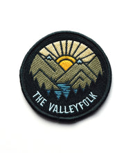 A black patch with a sun rising behind vector drawin mountains with a lake and silhouetted trees and “The Valleyfolk” in sans serif font at the bottom - from the Valleyfolk