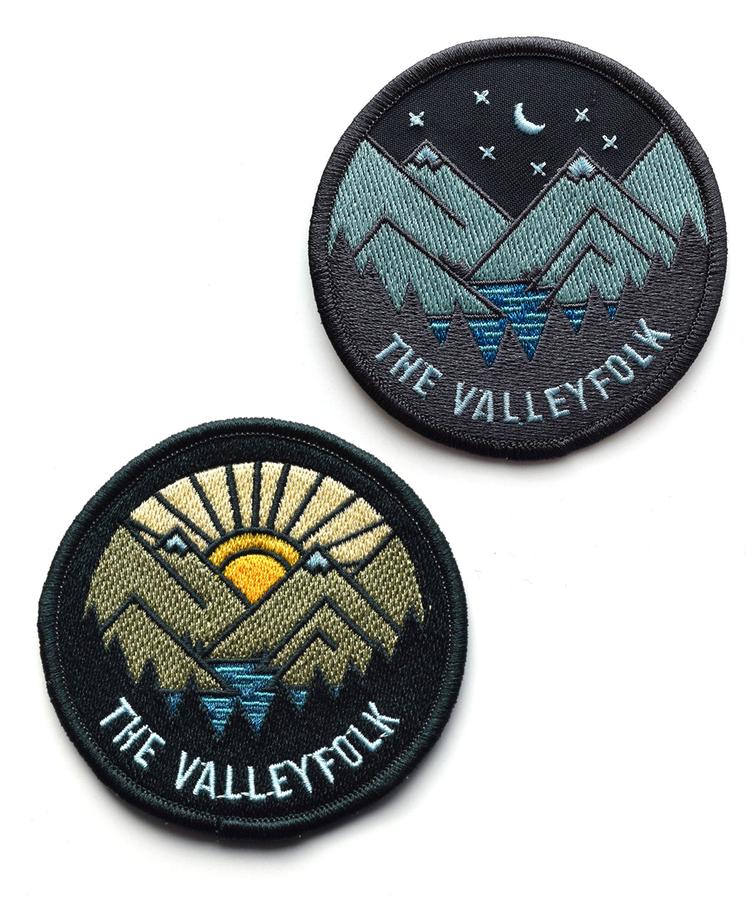 Two patches, one with a sun rising behind vector drawin mountains with a lake and silhouetted trees and “The Valleyfolk” in sans serif font at the bottom. The other patch is the same mountain scene at night - from the Valleyfolk