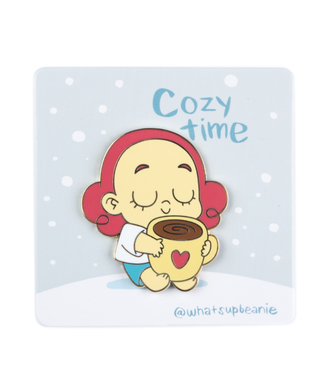 A pin of Beanie closing their eyes, with a smile holding a big mug of coffee with a heart on the front of the mug. The pin is on a card backing with falling snow and the words "Cozy Time; @whatsupbeanie". 