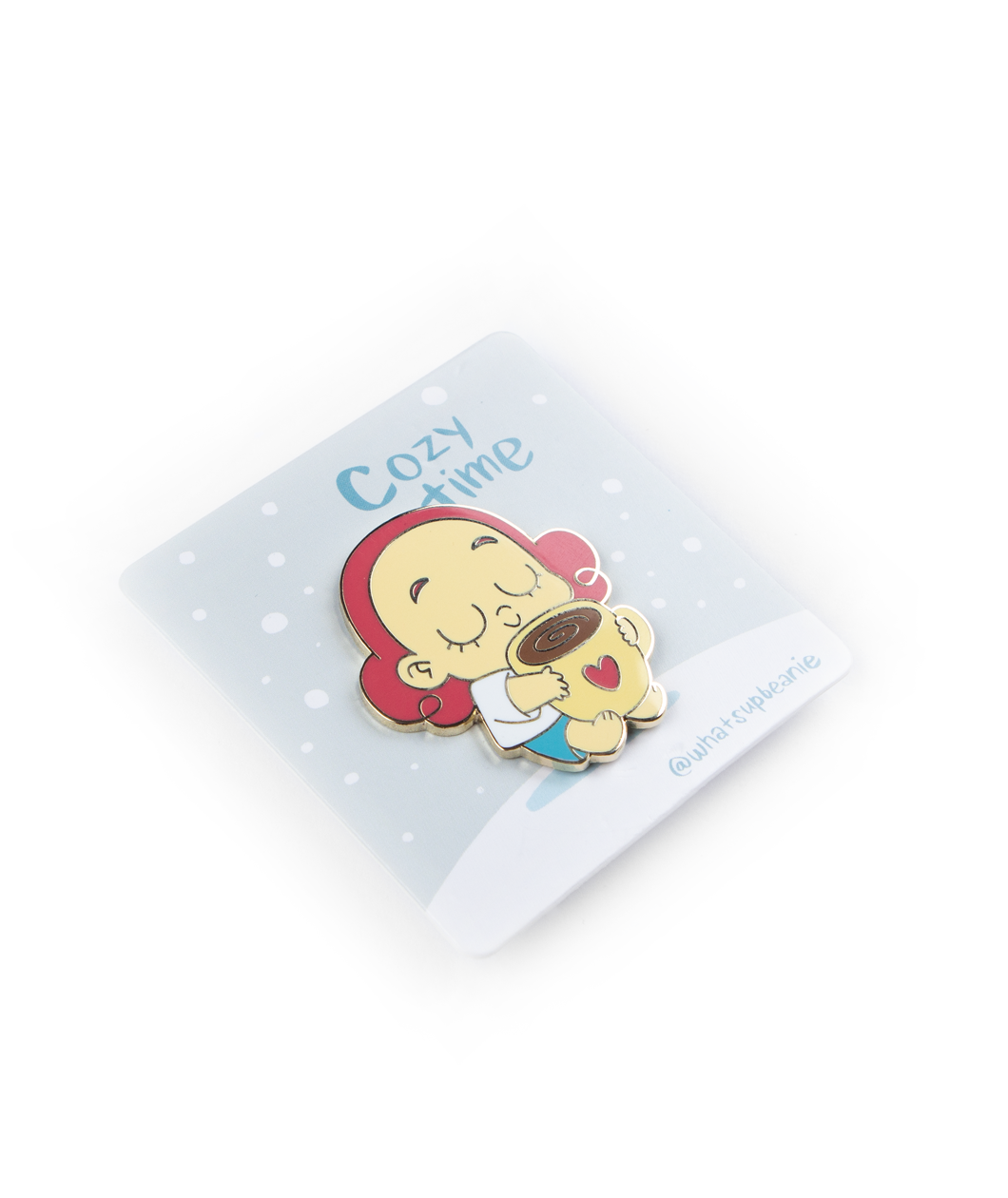  A pin of Beanie closing their eyes, with a smile holding a big mug of coffee with a heart on the front of the mug. The pin is on a card backing with falling snow and the words "Cozy Time; @whatsupbeanie".